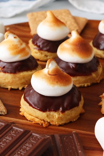 A wood tray of four s'mores cookies topped with chocolate and toasted meringue. Squares of Hershey's chocolates are in the forefront.
