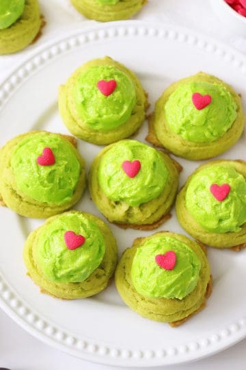 A round white plate of seven Grinch drop cookies topped with lime green frosting and a red heart sprinkle.