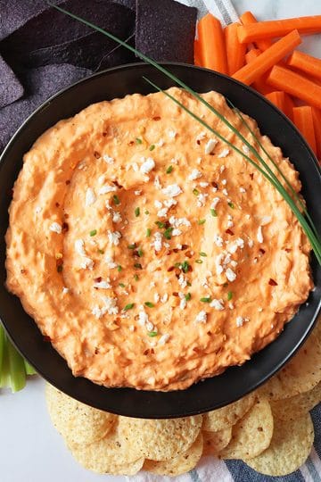 An overhead view of a round black bowl of buffalo chicken dip. Two stalks of chives lay over the bowl. Carrot sticks, celery stalks, blue chips, and round tortilla chips surround the bowl.